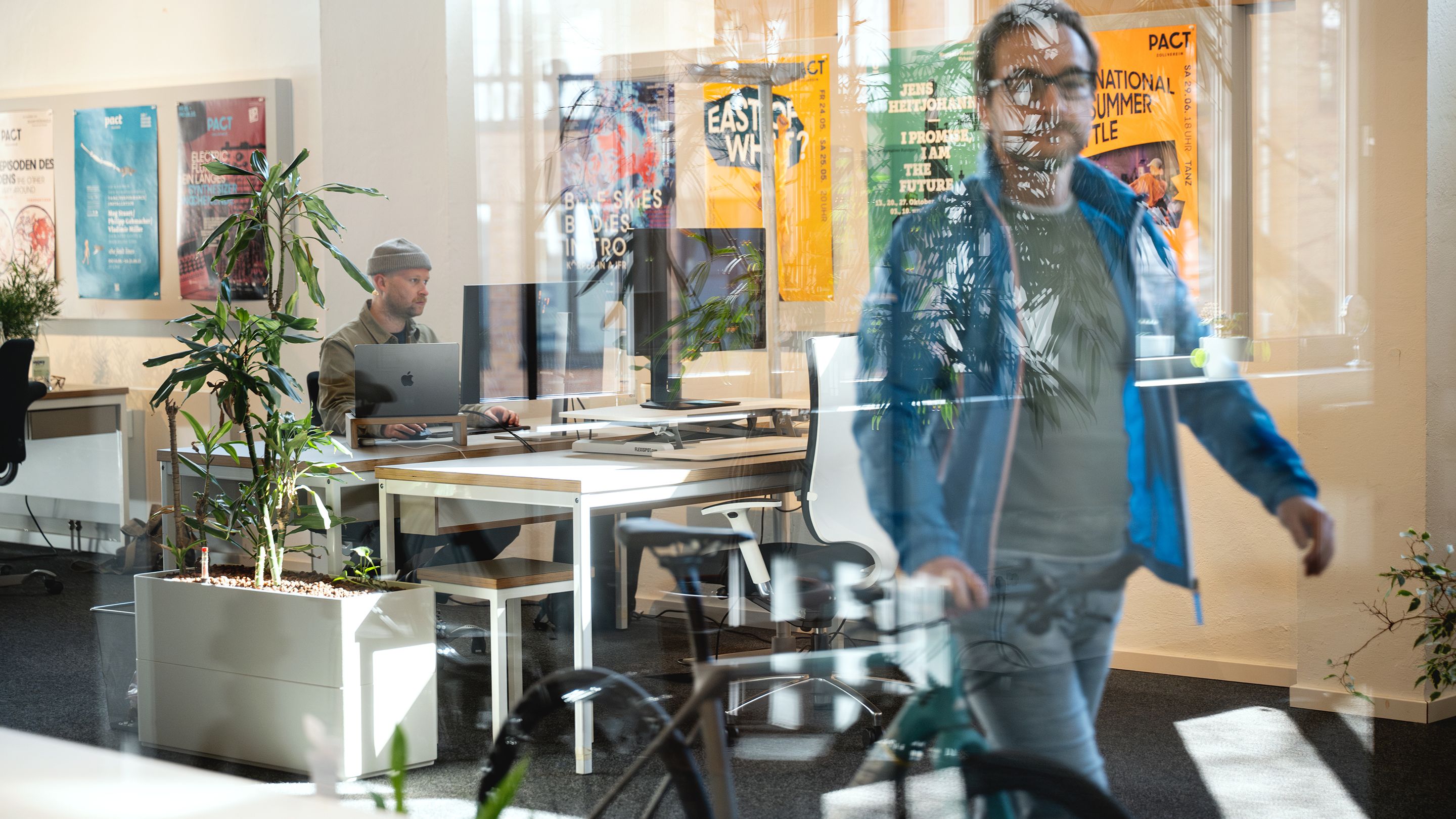 Ein Mitarbeitender von Labor b schiebt ein Rennrad durch das Büro. Im Hintergrund sitzt ein weiterer Mitarbeitender an seinem Schreibtisch und arbeitet.