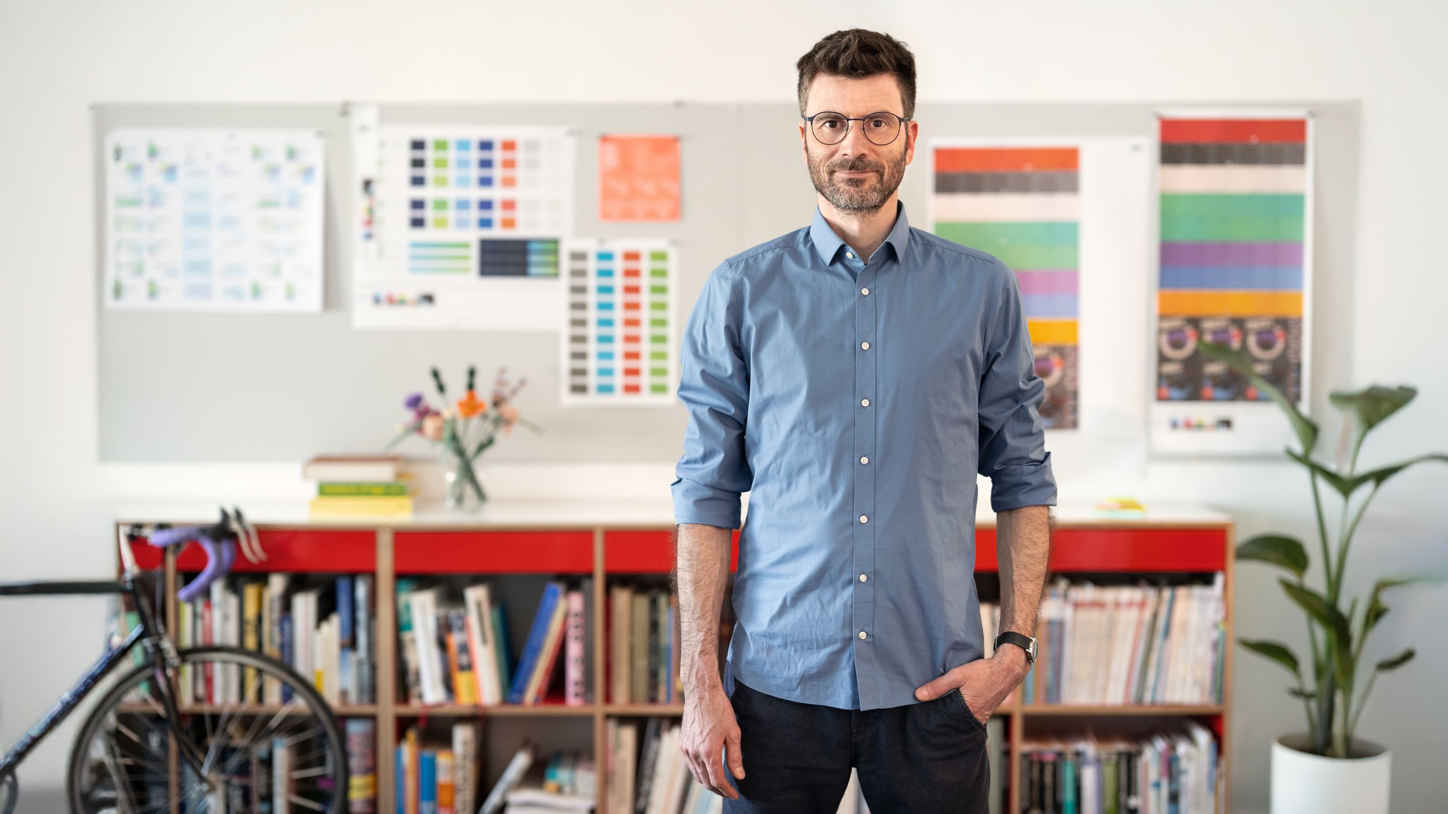 Labor b-Geschäftsführer Simon Busse steht erwartungsvoll vor einer Magnetwand mit Design-Entwürfen. Leicht unscharf zu erkennen sind außerdem ein Rennrad, ein Bücherregal und eine Topfpflanze.